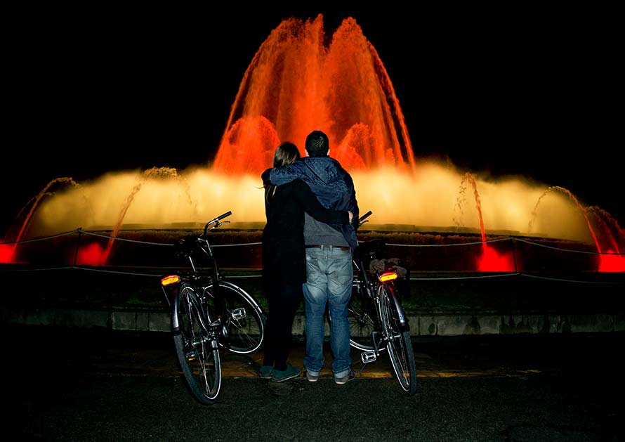 Two people standing next to their bikes enjoying an evening fountain water show